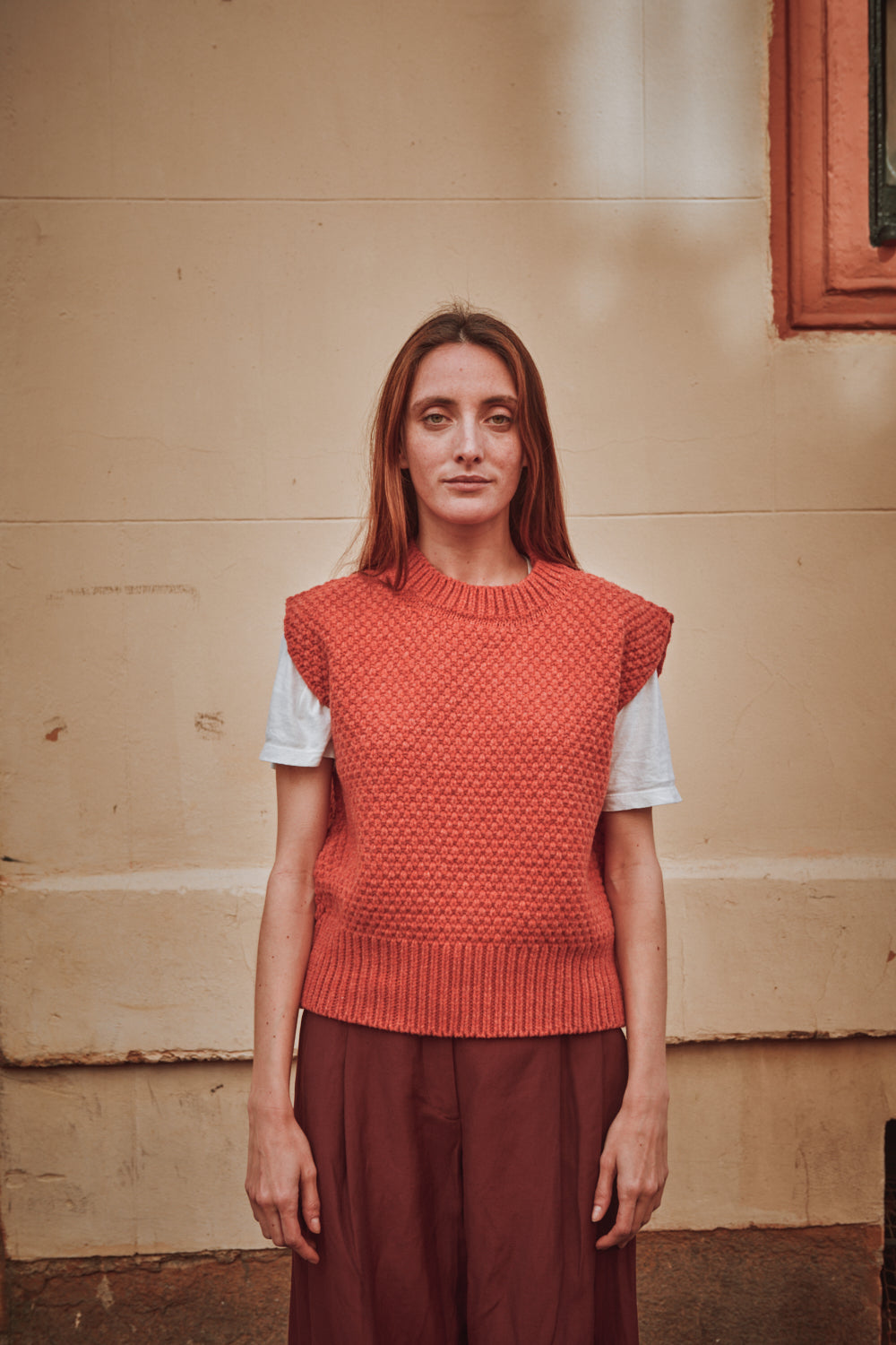 Femme portant un gilet en maille L'ENVERS corail sur une chemise blanche devant un mur beige.