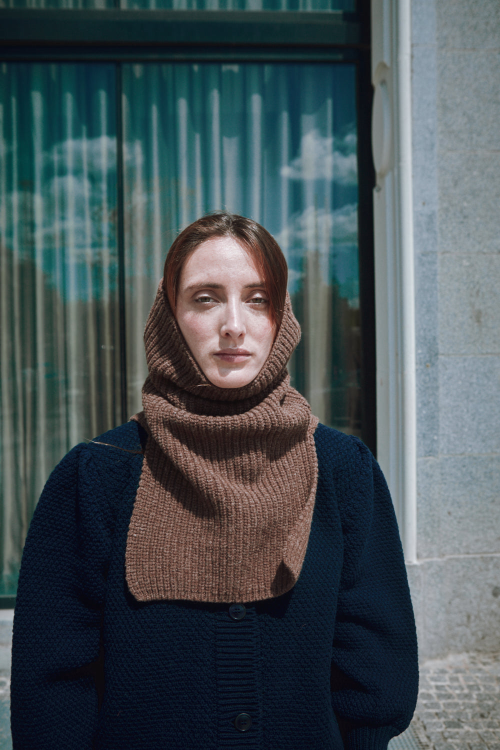 Femme portant une cagoule en tricot marron devant un bâtiment aux volets métalliques verts.