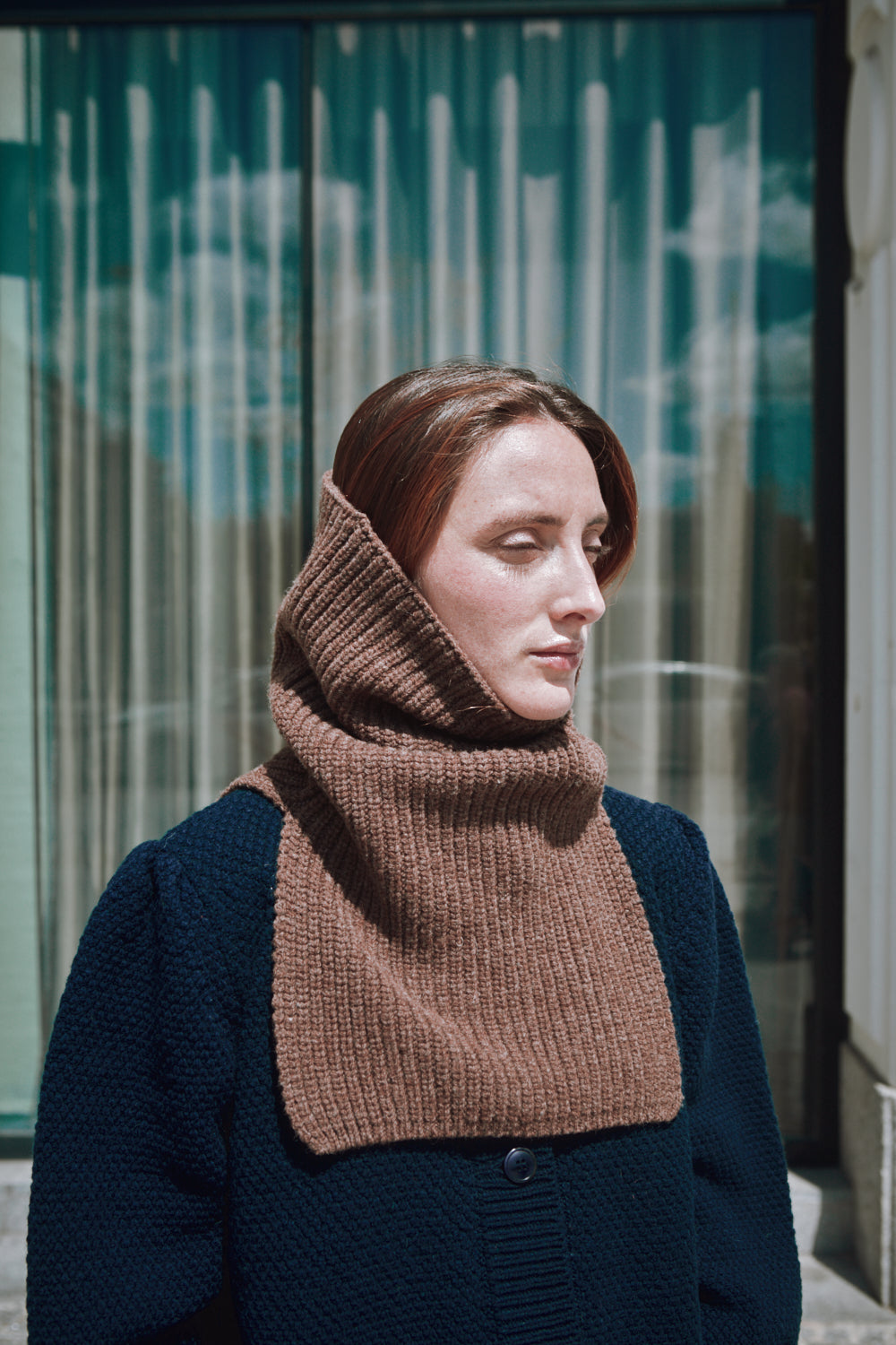 Femme portant une cagoule en tricot marron devant un bâtiment moderne.