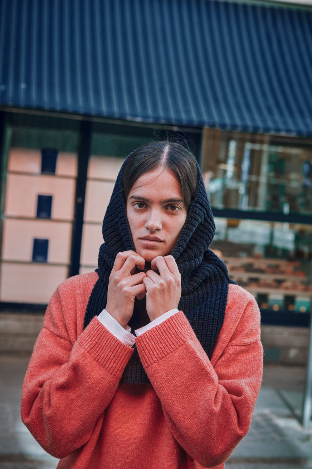 Personne portant un pull corail et une cagoule marine devant un bâtiment