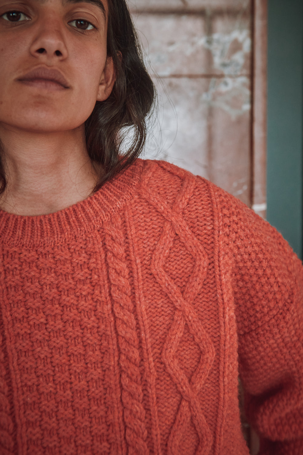 Personne portant un pull L'ENVERS orange corail et un pantalon blanc, debout dans une pièce aux portes vertes et au mur de marbre.