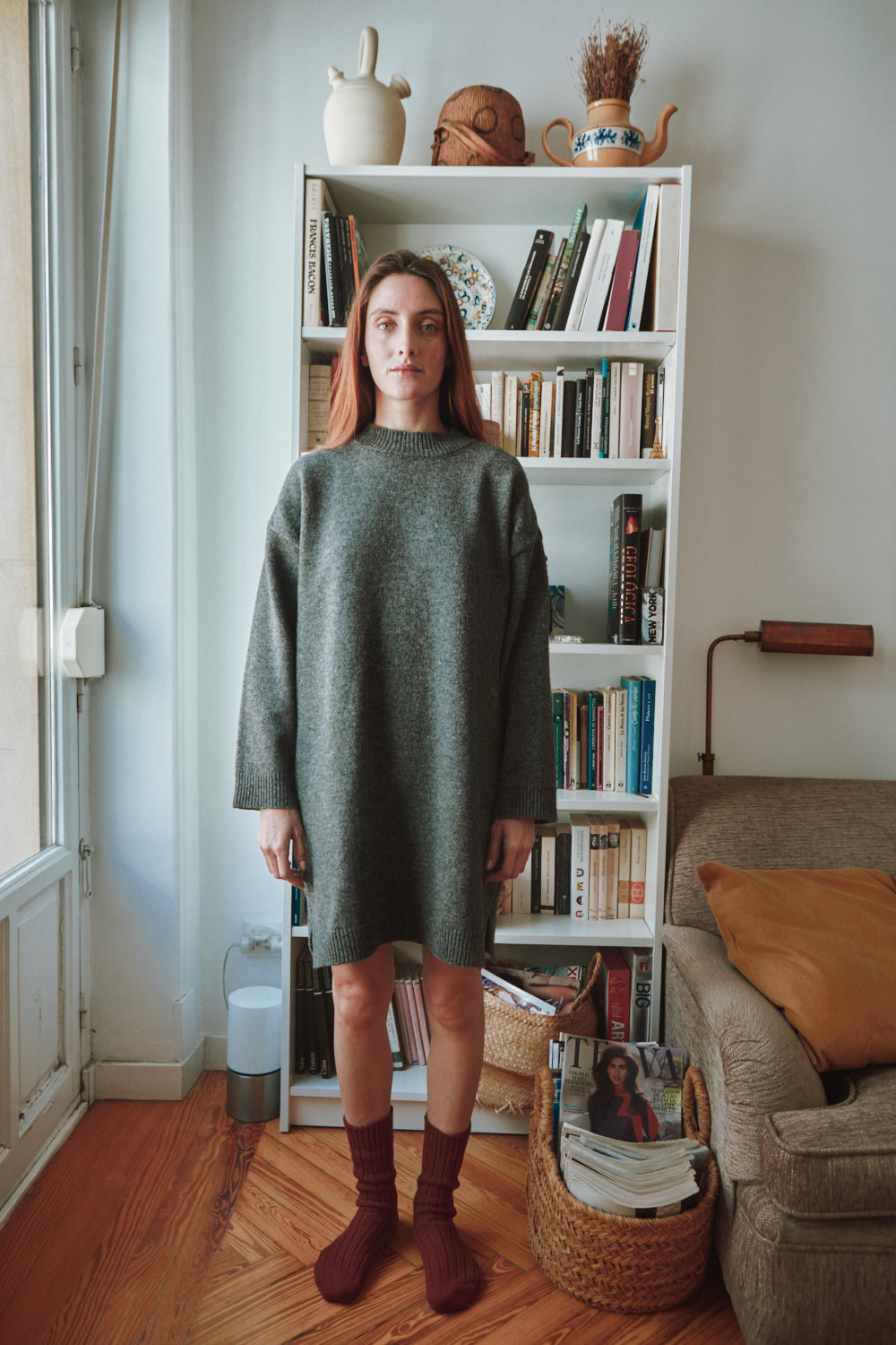 Femme en robe grise debout dans une pièce avec une bibliothèque et un canapé.