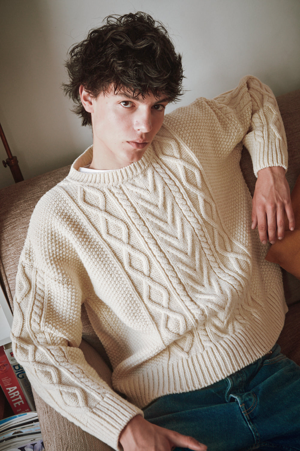 Personne portant un pull en tricot câblé crème, assise sur un canapé.