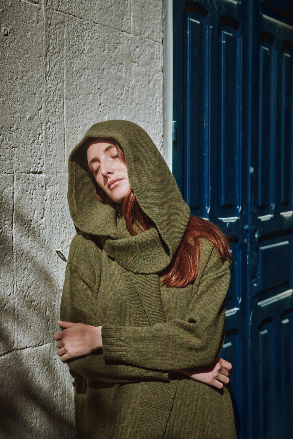 Personne portant une cagoule verte avec un pull-over assorti contre un mur texturé et une porte bleue.