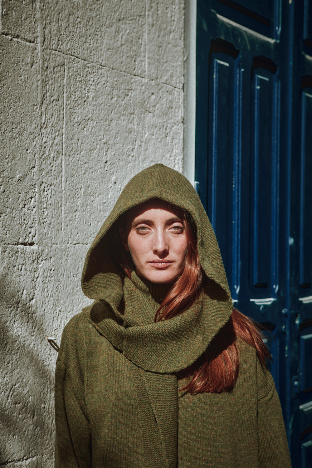 Personne portant une cagoule verte avec un pull-over assorti contre un mur texturé et une porte bleue.