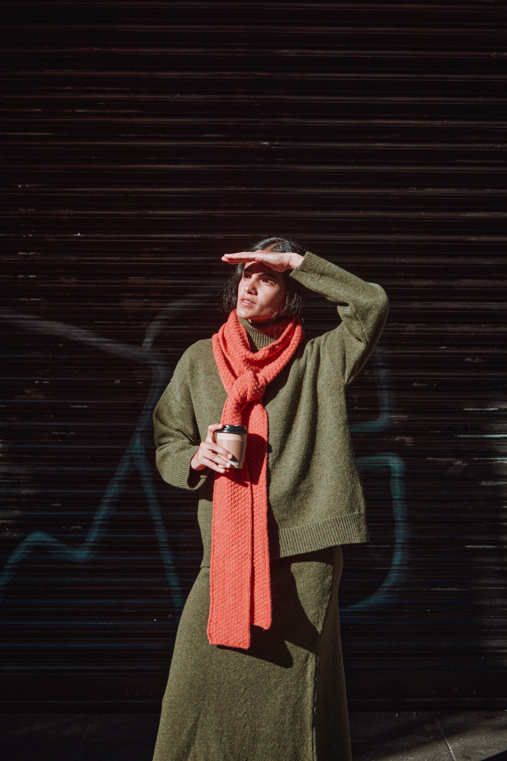 Personne portant un pull-over vert et une écharpe corail, debout sur un fond sombre