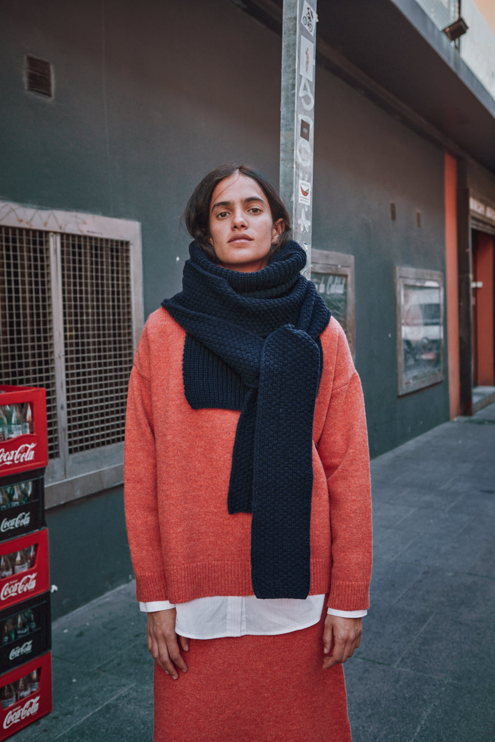 Personne portant un pull-over orange et une écharpe sombre, debout à côté de caisses de Coca-Cola contre un mur.
