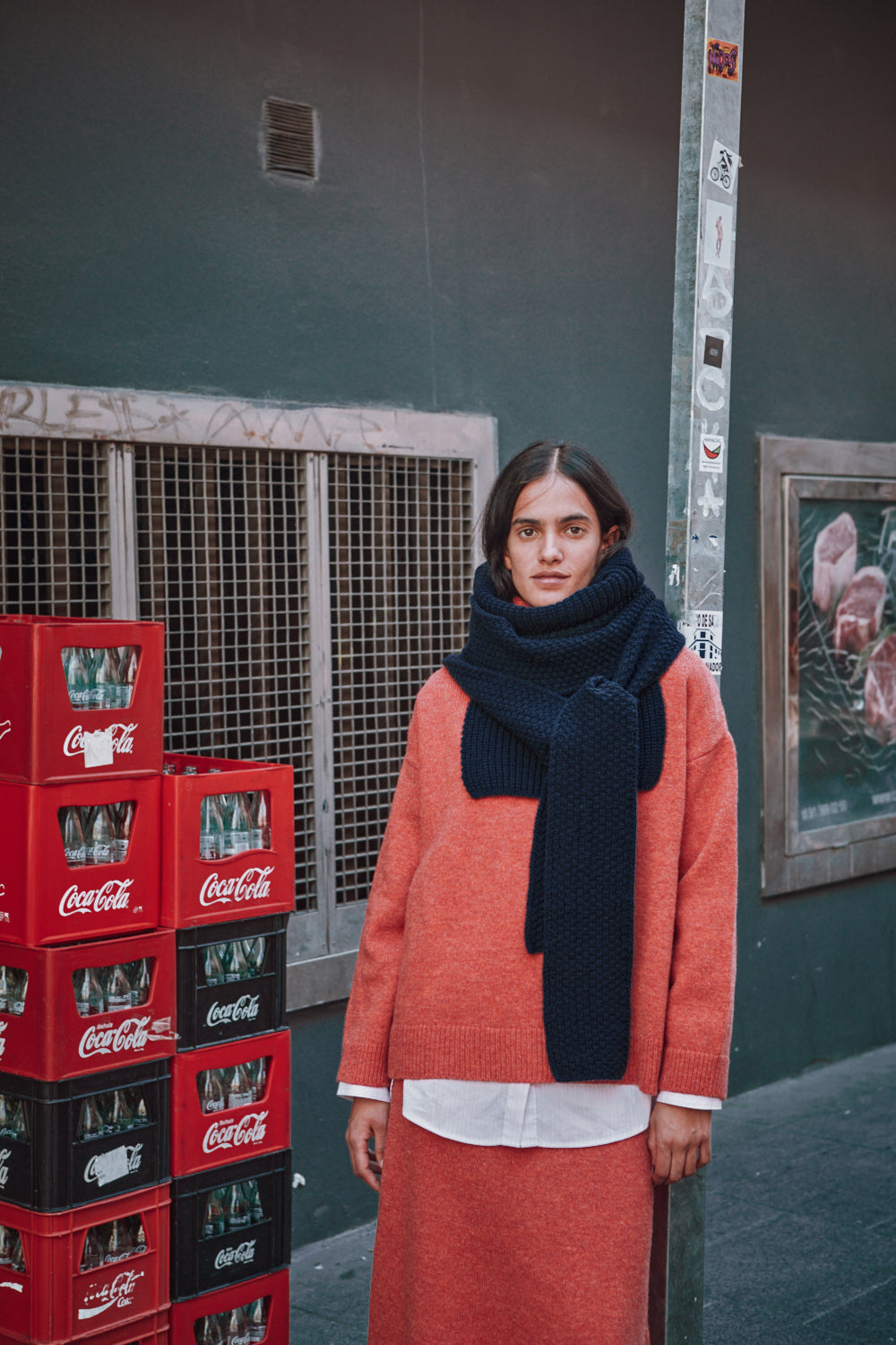 Personne portant un pull-over orange et une écharpe sombre, debout à côté de caisses de Coca-Cola contre un mur.