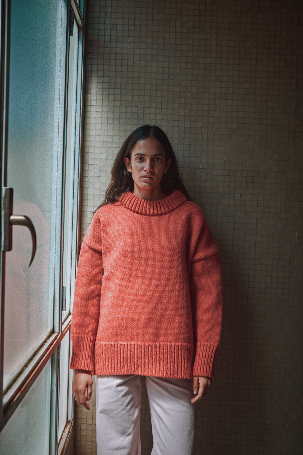 Personne portant un pull corail debout près d'une fenêtre avec un mur carrelé.