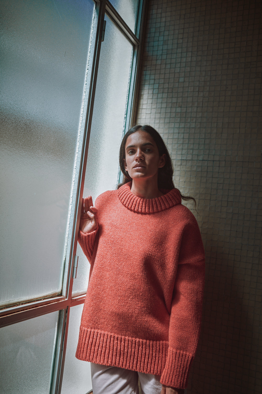 Personne portant un pull corail debout près d'une fenêtre avec un mur texturé.