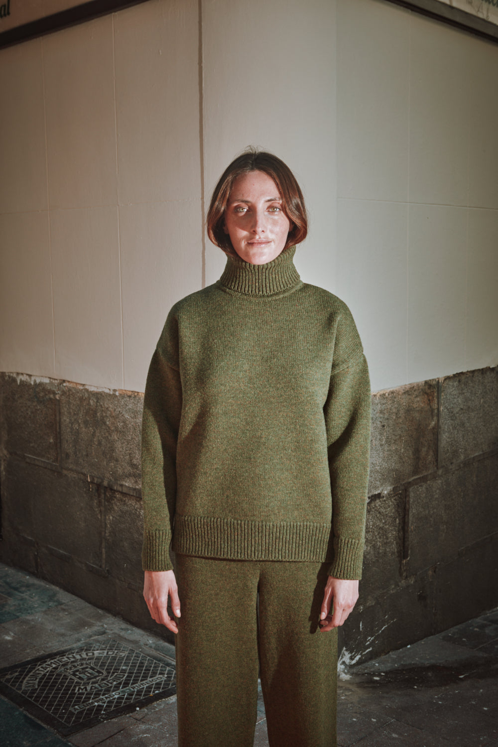Personne portant un pull à col roulé vert et un pantalon vert dans une pièce avec un mur en béton.