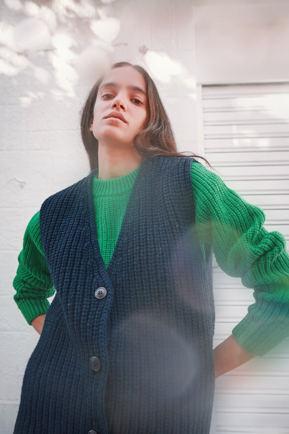 Personne portant un pull vert et un gilet bleu foncé contre un mur de briques blanches.