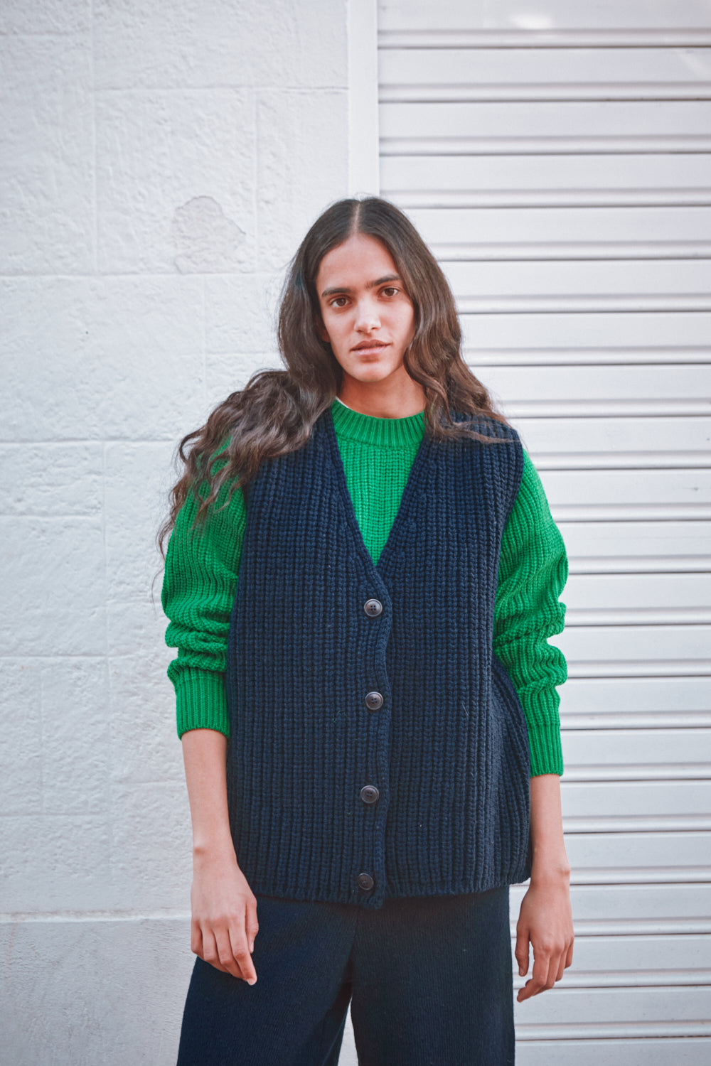 Femme portant un pull vert et un gilet bleu marine contre un mur blanc.